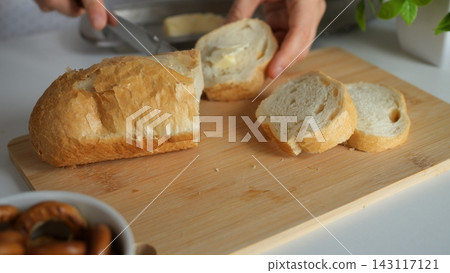 Person spreads creamy butter on sliced baguette over wooden cutting board during breakfast prep, demonstrating careful culinary knife skills while slicing fresh crusty French bread loaf. Person spreads creamy butter on sliced baguette over wooden cutting board during breakfast prep, demonstrating careful culinary knife skills while slicing fresh crusty French bread loaf. 143117121