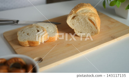 Crusty white bread loaf lies sliced on rectangular wooden board beside kitchen knife. Fresh bakery pastry demonstrates clean food preparation for morning meal atop smooth white table setting. Crusty white bread loaf lies sliced on rectangular wooden board beside kitchen knife. Fresh bakery pastry demonstrates clean food preparation for morning meal atop smooth white table setting. 143117135