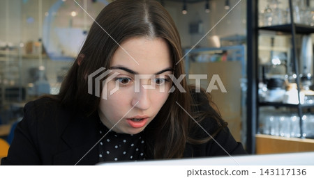Young woman gazes at laptop screen with astonished facial expression indoors. Surprised female professional reacts to unexpected digital notification while working inside modern coffee shop. Young woman gazes at laptop screen with astonished facial expression indoors. Surprised female professional reacts to unexpected digital notification while working inside modern coffee shop. 143117136