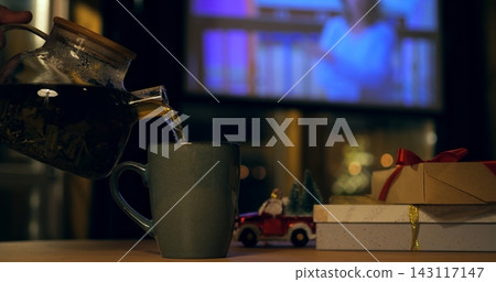Person pours hot tea from teapot into ceramic cup during winter tea ritual, offering warm comfort near Christmas gift boxes and miniature holiday car decor in dim evening interior lighting. Person pours hot tea from teapot into ceramic cup during winter tea ritual, offering warm comfort near Christmas gift boxes and miniature holiday car decor in dim evening interior lighting. 143117147