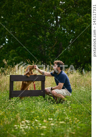 Young man kneeling beside dog during outdoor obstacle training session Young man kneeling beside dog during outdoor obstacle training session 143117331