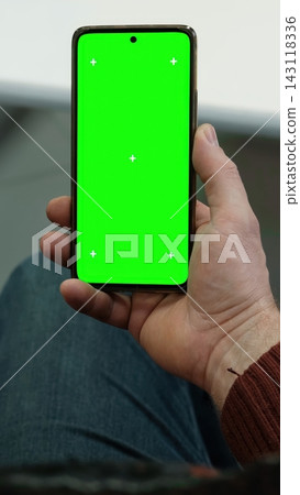 Man hand holding smartphone vertically with green screen chroma key and tracking markers over jeans. Close up view of male palm with isolated cellular template mockup inside office. Vertical photo. Man hand holding smartphone vertically with green screen chroma key and tracking markers over jeans. Close up view of male palm with isolated cellular template mockup inside office. Vertical photo. 143118336