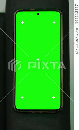 Isolated green screen chroma key artificial smartphone mockup display with tracking markers lying on a black fabric couch armrest. Static flat lay picture of phone template backdrop. Vertical shot. Isolated green screen chroma key artificial smartphone mockup display with tracking markers lying on a black fabric couch armrest. Static flat lay picture of phone template backdrop. Vertical shot. 143118337