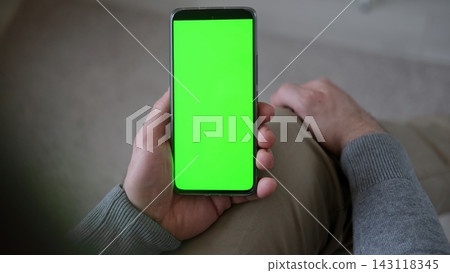 Top view of man holding smartphone vertically with green screen mockup chroma key blank display. Male user sitting and looking at mobile phone template mockup surface inside cozy room at home Top view of man holding smartphone vertically with green screen mockup chroma key blank display. Male user sitting and looking at mobile phone template mockup surface inside cozy room at home 143118345