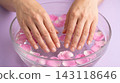 Close-up of a woman's hands soaking in a clear glass bowl of warm water with pink flower petals for a relaxing manicure prep. 143118646