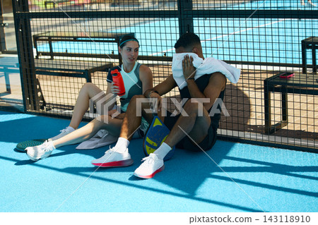 Male and female padel players relaxing on court during break from outdoor exercise Male and female padel players relaxing on court during break from outdoor exercise 143118910