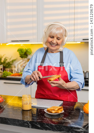 Senior old woman dancing with headphones while making peanut butter toast in home kitchen for diet Senior old woman dancing with headphones while making peanut butter toast in home kitchen for diet 143120679