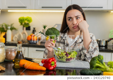 Young woman tired of keto diet eats fresh vegetable salad in modern kitchen, feeling discouraged now Young woman tired of keto diet eats fresh vegetable salad in modern kitchen, feeling discouraged now 143120740