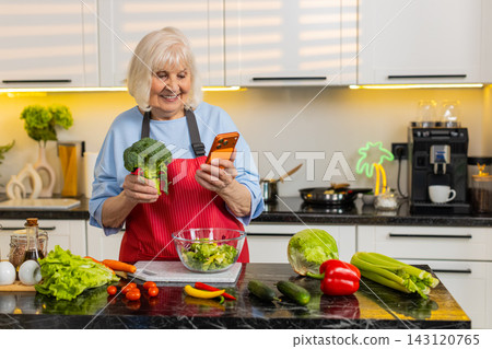 Senior old woman checking smartphone while making healthy vegetable salad in home kitchen diet plan Senior old woman checking smartphone while making healthy vegetable salad in home kitchen diet plan 143120765
