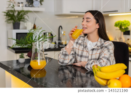 Young woman drinks fresh orange juice from glass, enjoying healthy hydration in home kitchen 143121190