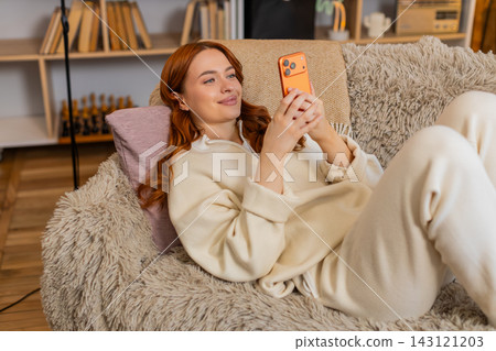 Young woman on home sofa smiling with smartphone while resting and scrolling during leisure indoors Young woman on home sofa smiling with smartphone while resting and scrolling during leisure indoors 143121203