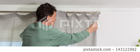A man is hanging a curtain on the rod after washing it. It is spring, and he is doing household chores in a room with a window and some greenery, banner A man is hanging a curtain on the rod after washing it. It is spring, and he is doing household chores in a room with a window and some greenery, banner 143122992