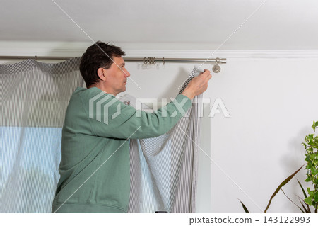 A man is hanging a curtain on the rod after washing it. It is spring, and he is doing household chores in a room with a window and some greenery A man is hanging a curtain on the rod after washing it. It is spring, and he is doing household chores in a room with a window and some greenery 143122993
