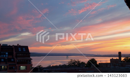 Pink and orange clouds illuminate the evening sky above a coastal city and calm sea framed by dark silhouettes. The scene expresses harmony between urban life and nature during the quiet transition 143123083