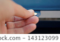 A hand holds a tiny metallic jewelry component in front of a blue storage case. The close-up detail represents precision, craftsmanship, and careful handling of small accessory parts. 143123099