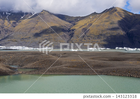 Icebergs beyond gravel shore and turquoise glacial pond beneath mountains in Iceland Icebergs beyond gravel shore and turquoise glacial pond beneath mountains in Iceland 143123318