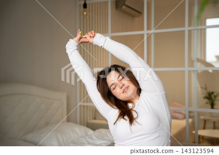 Young woman stretching with raised arms beside bed in a bright modern bedroom. Slow morning wellness self-care and healthy lifestyle 143123594