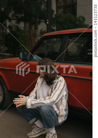 Guy with long hair squatting next to red car on city street during daytime in urban area Guy with long hair squatting next to red car on city street during daytime in urban area 143123738