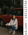Guy with long hair sits by red car on city street during daylight hours 143123744