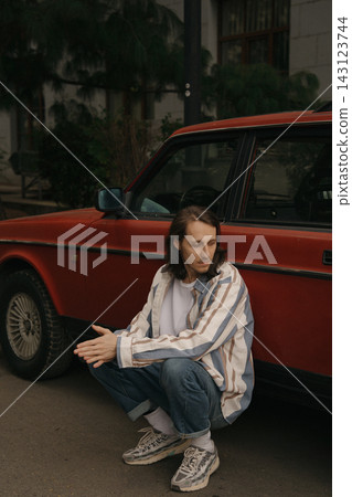 Guy with long hair sits by red car on city street during daylight hours Guy with long hair sits by red car on city street during daylight hours 143123744