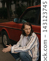 Guy with long hair sits by a red car in an urban area during daytime 143123745