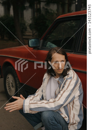 Guy with long hair sits by a red car in an urban area during daytime 143123745