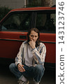 Guy with long hair squatting by red car during daytime in urban area 143123746