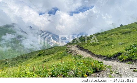Mountain trail climbs through Svaneti, wide view across green alpine slopes toward cloud-covered Caucasus peaks Mountain trail climbs through Svaneti, wide view across green alpine slopes toward cloud-covered Caucasus peaks 143123888