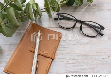 畫面中，白色木桌上放著一本皮質筆記本、一副黑框眼鏡和一支鋼筆。 143124616