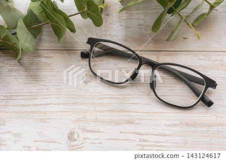一副時尚的黑框眼鏡和一幅桉樹風景畫擺放在一張白色木桌上。 一副時尚的黑框眼鏡和一幅桉樹風景畫擺放在一張白色木桌上。 143124617