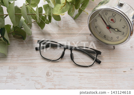 一副時尚的黑框眼鏡、一個模擬時鐘和一幅植物風景畫擺放在一張白色木桌上。 一副時尚的黑框眼鏡、一個模擬時鐘和一幅植物風景畫擺放在一張白色木桌上。 143124618
