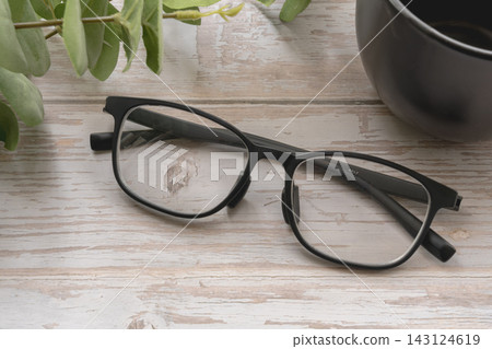 一副時尚的黑框眼鏡、一個黑色馬克杯和一幅植物風景畫擺放在一張白色木桌上。 一副時尚的黑框眼鏡、一個黑色馬克杯和一幅植物風景畫擺放在一張白色木桌上。 143124619