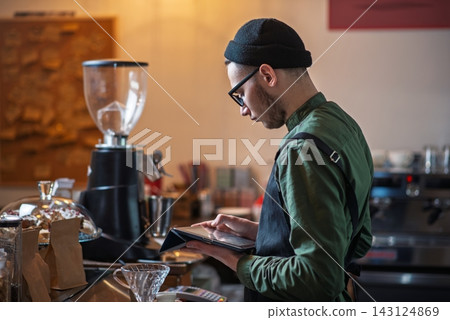 Young male barista using a digital tablet at a cafe counter Young male barista using a digital tablet at a cafe counter 143124869