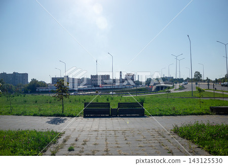 Wooden park benches on paved walkway facing city industrial zone under bright sunny sky for urban infrastructure development 143125530