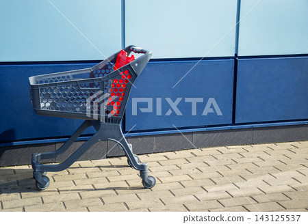 Empty grey plastic shopping cart with red details standing on paved sidewalk by blue wall outdoors with copy space for retail store concept Empty grey plastic shopping cart with red details standing on paved sidewalk by blue wall outdoors with copy space for retail store concept 143125537