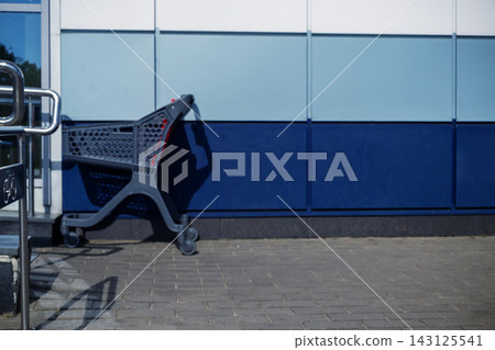Empty plastic shopping trolley on paved sidewalk near blue wall with copy space outdoors for supermarket retail concepts Empty plastic shopping trolley on paved sidewalk near blue wall with copy space outdoors for supermarket retail concepts 143125541