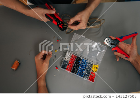 Workers hands precisely crimp electrical wire terminals with crimping tool, colorful connectors laid out on gray surface for precise installation and maintenance work, close-up, cropped shot Workers hands precisely crimp electrical wire terminals with crimping tool, colorful connectors laid out on gray surface for precise installation and maintenance work, close-up, cropped shot 143127503