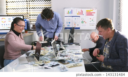 Four employees are counting money from a major investor. A group of people are sitting at a table with laptops and dollars in cash 143131484