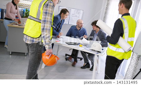 A group of workers and architects stands around a table with drawings and building plans. Discussion of the project budget. A meeting or briefing 143131495