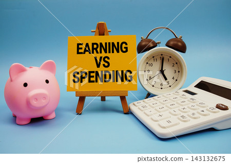 Earning Vs Spending text with piggy bank, alarm clock and calculator on blue background Earning Vs Spending text with piggy bank, alarm clock and calculator on blue background 143132675