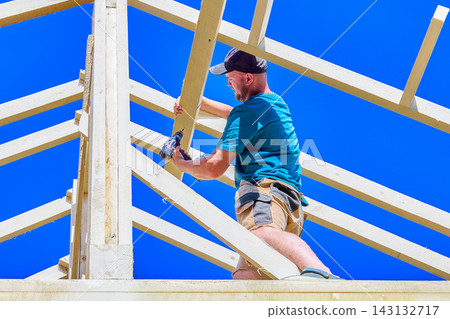 Rafters form timber roof framework as young construction worker drives a screw with cordless drill during assembly. Rafters form timber roof framework as young construction worker drives a screw with cordless drill during assembly. 143132717