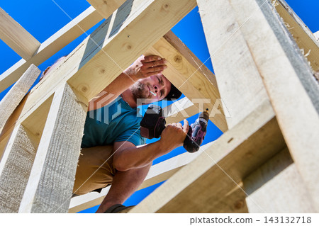 Roof truss connects wooden rafter legs while builder fastens timber joint using battery powered screwdriver. Roof truss connects wooden rafter legs while builder fastens timber joint using battery powered screwdriver. 143132718
