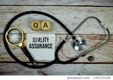 Quality Assurance (QA) text with stethoscope and magnifying glass on wooden background Quality Assurance (QA) text with stethoscope and magnifying glass on wooden background 143133109