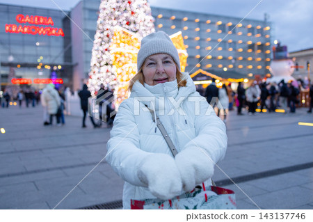 Senior female tourist holding a festive holiday gift bag and smiling in front of a decorated Christmas tree on a European city square during New Year 2027 celebration. Senior female tourist holding a festive holiday gift bag and smiling in front of a decorated Christmas tree on a European city square during New Year 2027 celebration. 143137746
