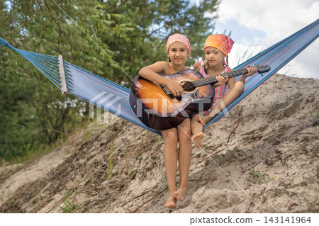 The girls are sitting in a hammock on the beach, playing guitar together The girls are sitting in a hammock on the beach, playing guitar together 143141964