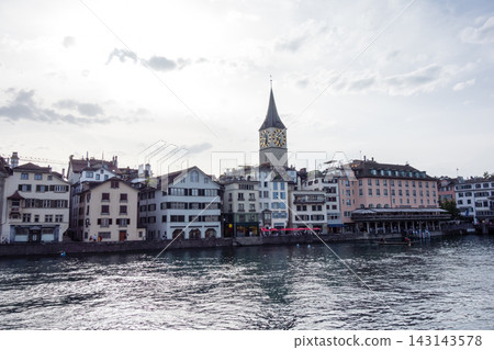 A church with a large clock tower and colorful buildings stand along the river in the bright sunlight – the cityscape of Zurich, Switzerland. A church with a large clock tower and colorful buildings stand along the river in the bright sunlight – the cityscape of Zurich, Switzerland. 143143578