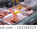 A shopper's hands using tongs to select a salmon fillet in the fresh fish section of a supermarket. 143144319