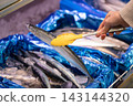 A shopper's hands using tongs to select fresh saury (Pacific saury) in the fresh fish section of a supermarket. 143144320
