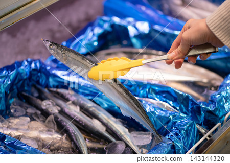 顧客用夾子在超市的鮮魚區挑選新鮮的秋刀魚（太平洋秋刀魚）。 143144320