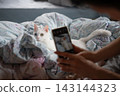 A pet owner's daily routine: taking photos of their white cat relaxing in bed with their smartphone. 143144323